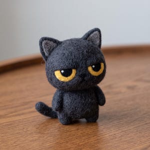 Op de afbeelding: Een klein, naaldgevilt donkergrijs kattenfiguurtje met een textuurrijk, pluizig oppervlak. De kat heeft grote, opvallende gele ogen met zwarte pupillen, puntige oren met lichtgrijze details aan de binnenkant en een kleine, gebogen staart.