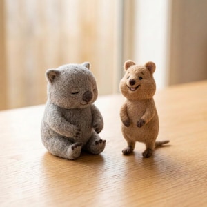 May include: Two needle-felted animal figurines. On the left, a gray wombat sits with eyes closed. On the right, a light brown quokka stands upright with a smiling expression, dark eyes, and a small tail. Both figures have textured wool surfaces.