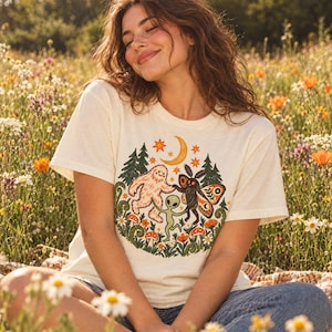 Può includere: Una t-shirt a maniche corte color crema con una stampa grafica colorata che raffigura un sasquatch, un alieno e una falena che ballano insieme in un ambiente boschivo con funghi, felci, pini, una luna crescente e stelle.