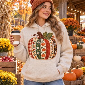 May include: A light beige hooded sweatshirt featuring a large, central graphic of a pumpkin composed of vertical panels with varied patterns, including red plaid, floral motifs, polka dots, and small orange pumpkin illustrations.