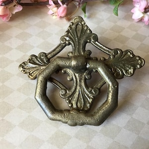 May include: Antique brass drawer pull with an ornate design. The pull features a hexagonal handle and decorative flourishes. The hardware has a vintage aesthetic, with a slightly aged appearance. The background includes pink flowers and a patterned surface.