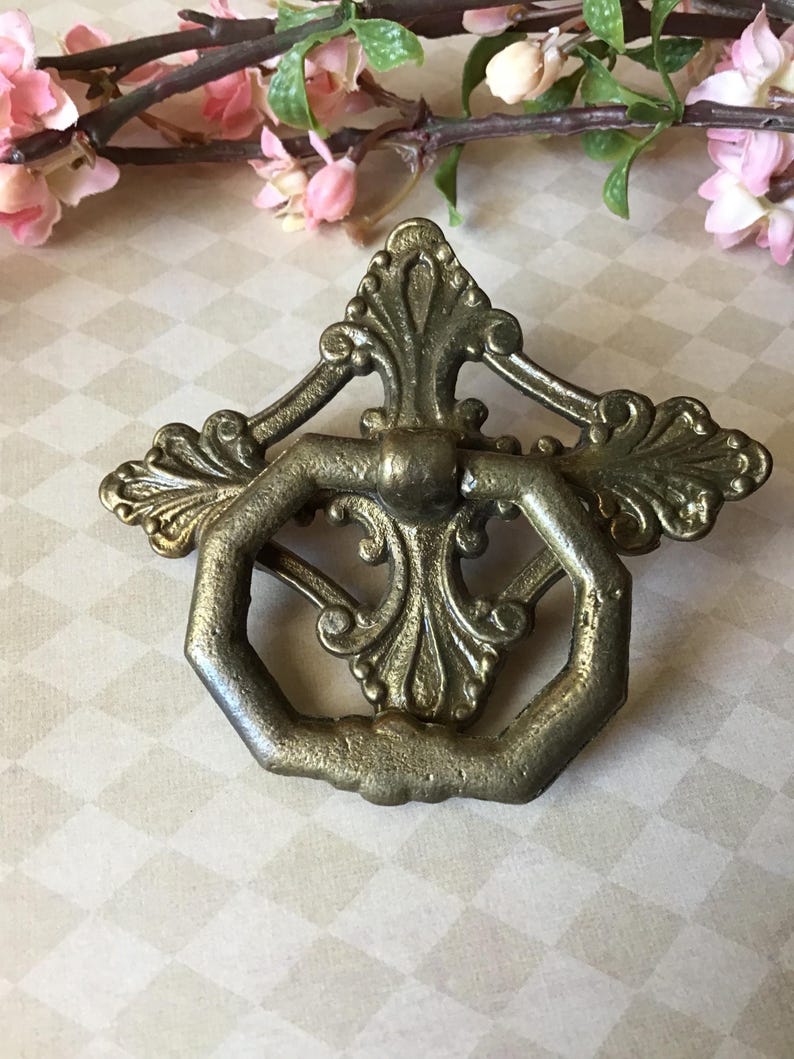 Peut inclure: Poign&eacute;e de tiroir en laiton antique avec un design orn&eacute;. La poign&eacute;e pr&eacute;sente une poign&eacute;e hexagonale et des ornements d&eacute;coratifs. Le mat&eacute;riel a une esth&eacute;tique vintage, avec une apparence l&eacute;g&egrave;rement vieillie. Le fond comprend des fleurs roses et une surface &agrave; motifs.