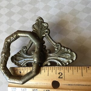 Peut inclure: Tirette de tiroir en laiton antique avec des d&eacute;tails floraux et des volutes orn&eacute;s. La tirette a une poign&eacute;e hexagonale et une plaque arri&egrave;re d&eacute;corative. La r&egrave;gle indique une longueur d'environ 5 cm.