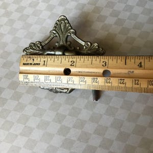 Peut inclure: Poign&eacute;e de tiroir antique en laiton orn&eacute; de volutes d&eacute;coratives. La poign&eacute;e mesure environ 7,6 cm de long. Comprend une vis de montage. Parfait pour ajouter une touche vintage aux meubles.