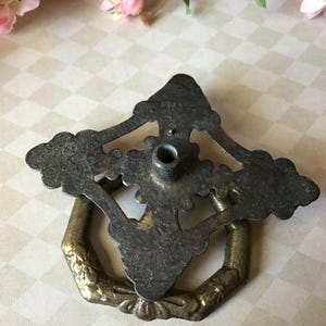 Peut inclure: Tirette de tiroir en laiton antique avec un design orn&eacute;. La tirette pr&eacute;sente une finition sombre et patin&eacute;e sur la plaque arri&egrave;re et une poign&eacute;e en anneau hexagonale. Le design comprend des d&eacute;tails floraux et des volutes, ajoutant une esth&eacute;tique vintage.