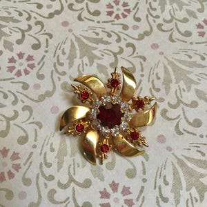 May include: A gold-toned brooch in a floral design, featuring a large, round, red gemstone center surrounded by smaller red and clear gemstones. The brooch has curved petal-like elements.