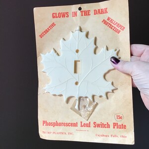May include: A white, leaf-shaped light switch plate on a vintage cardboard backing. The plate is phosphorescent and has a rectangular opening for the switch. Text on the packaging reads "GLOWS IN THE DARK" and "Phosphorescent Leaf Switch Plate."