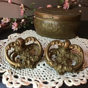 May include: Two antique brass drawer pulls with ornate floral designs sit on a white lace doily. A matching brass trinket box with a detailed lid is in the background. Pink flowers and branches add a touch of color to the scene.