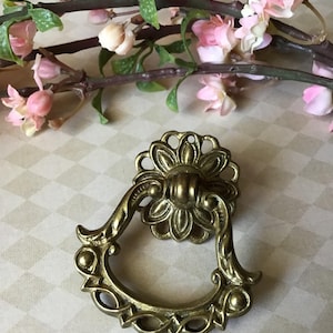 May include: Antique brass drawer pull with a floral design. The pull features a decorative flower at the top and a curved handle with intricate detailing. The hardware is a warm gold color, perfect for vintage furniture restoration.