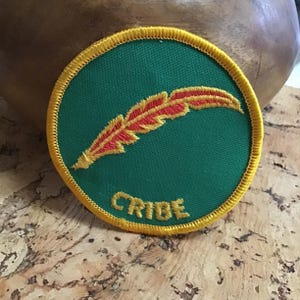 May include: A circular green patch with a gold border and the word "CRIBE" embroidered in gold. The patch features a red and gold feather design. This patch is likely for clothing or accessories.