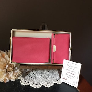May include: A vintage pink skai-hide vinyl wallet and card case set in a cream-colored box with gold trim. The set is displayed on a small wooden easel with a white lace doily. A small card reads "This fine product is made of exclusive, genuine skai-hide the luxury vinyl of our time."