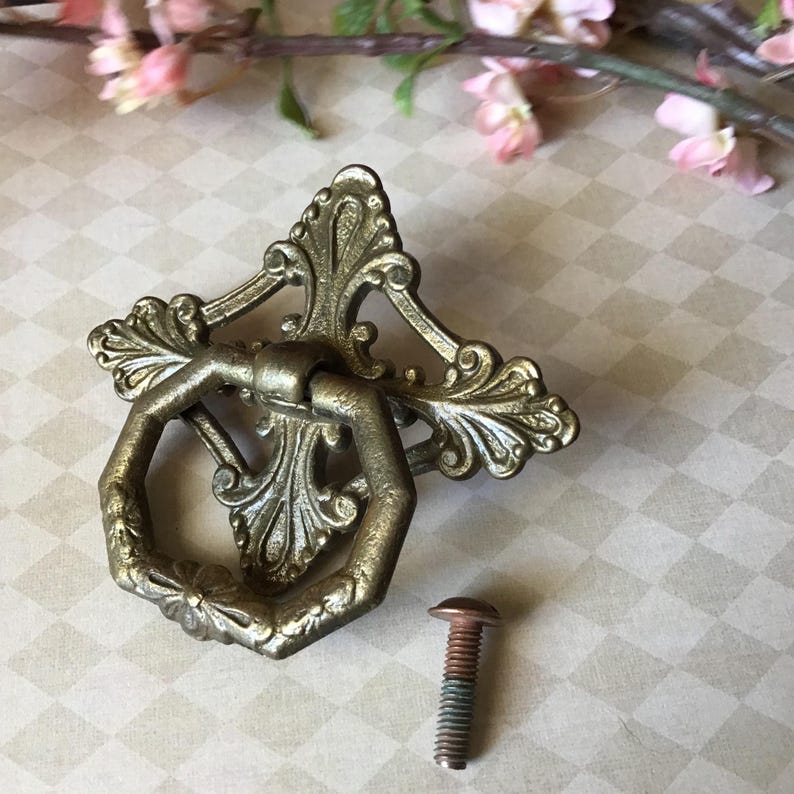 Peut inclure: Tirette de tiroir en laiton orn&eacute; avec une plaque arri&egrave;re d&eacute;corative et une poign&eacute;e hexagonale. Une vis assortie est incluse. La quincaillerie a une finition antique et est probablement destin&eacute;e &agrave; une armoire ou une commode. Le fond a un motif de losanges.