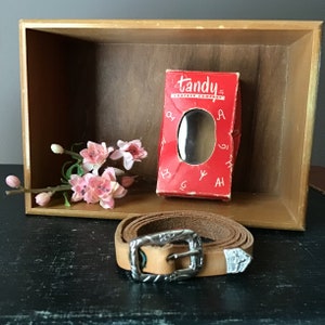 May include: A tan leather belt with a silver buckle and decorative tips. The belt is coiled on a black surface, next to a red box from Tandy Leather Company and a sprig of pink flowers. The box has the company name and logo.