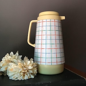 May include: A vintage yellow and white plaid thermos with a handle and lid. The thermos has a white background with a red, green, and blue plaid pattern. The base, handle, and lid are yellow. Decorative flowers are in the foreground.