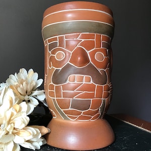 May include: A brown ceramic vase with a stylized face design. The vase features a cylindrical shape with a wide base and a detailed pattern of geometric shapes in shades of brown and tan. White accents highlight the design.