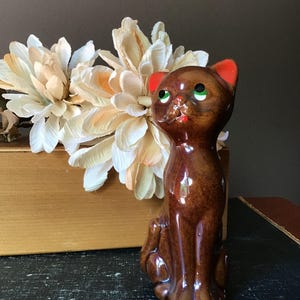 May include: A brown ceramic cat figurine with red ears and green eyes. The cat is sitting upright with a glossy finish. Artificial cream-colored flowers are in the background, adding a touch of elegance to the scene.