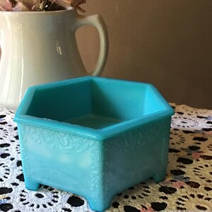 May include: A turquoise-colored, hexagonal trinket dish with a floral embossed design on the sides. The dish sits on small feet and is placed on a white lace doily. A white pitcher and dried flowers are in the background.
