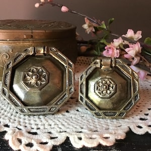May include: Two antique brass drawer pulls with an octagonal shape and a central floral design. The pulls have a hinged handle and are set against a backdrop of a lace doily and a decorative box.