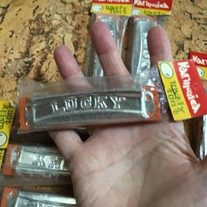 May include: A collection of silver harmonicas, each packaged in clear plastic. The word "LUCKY" is embossed on the front of one harmonica. The packaging includes orange and red accents with the text "Lovely Tone".