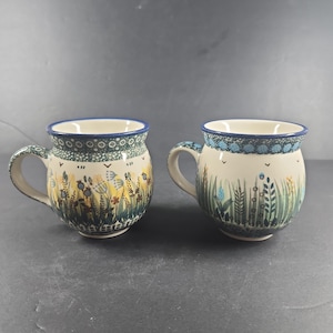May include: Two rounded ceramic mugs featuring a cream-coloured base with a hand-painted botanical pattern of green grasses, yellow flowers, and blue accents. Each mug has a blue rim and a curved handle, showcasing a distinct folk-art style design.