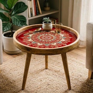 May include: A round wooden side table with three tapered legs and a red resin tabletop featuring a detailed, intricate mandala pattern in white, gold, and teal. A small succulent in a white pot and a glass sit on the table surface.
