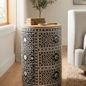 May include: A cylindrical bedside table featuring a black finish with intricate white floral and geometric inlay patterns. The piece includes three stacked drawers, each equipped with a small circular metal pull handle, and a natural wood top.