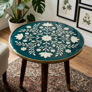 May include: A round side table with a deep teal resin top featuring an intricate white floral inlay pattern. The table has a thin brass-toned metal rim and three tapered dark wood legs with brass-toned metal caps at the bottom of each leg.