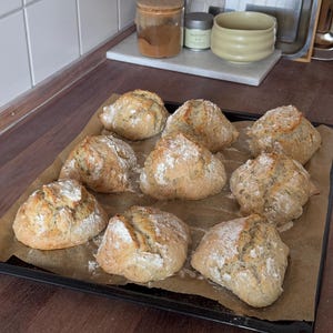 May include: Eight golden-brown, rustic bread rolls arranged on a black baking sheet lined with parchment paper. Each roll has a lightly floured, cracked crust with visible seeds throughout the dough, showcasing a rounded, uneven shape.
