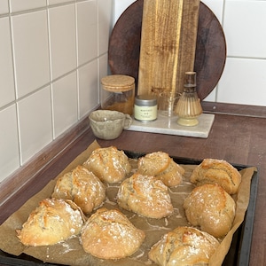Op de afbeelding: Een zwarte metalen bakplaat bevat negen goudbruine, met bloem bestoven broodjes, gerangschikt op bruin bakpapier. De broodjes hebben een rustieke, onregelmatige textuur met zichtbare barsten in de korst, wat duidt op een vers uiterlijk.