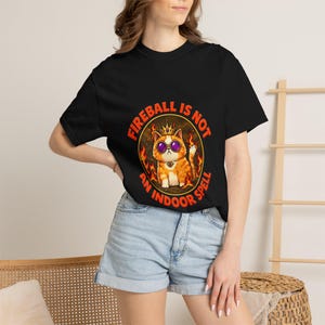 以下が含まれることがあります： 黒の半袖Tシャツ。金色の王冠と紫色のサングラスを身につけたオレンジ色の猫が炎に囲まれているグラフィックが描かれています。猫の周りにはオレンジ色の文字で「FIREBALL IS NOT AN INDOOR SPELL」と円状にプリントされています。