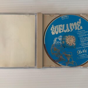 May include: An open CD case with a blue disc featuring the band name "Sublime" in white text. The disc also lists song titles and the "Skunk Records" logo. The case is clear plastic.