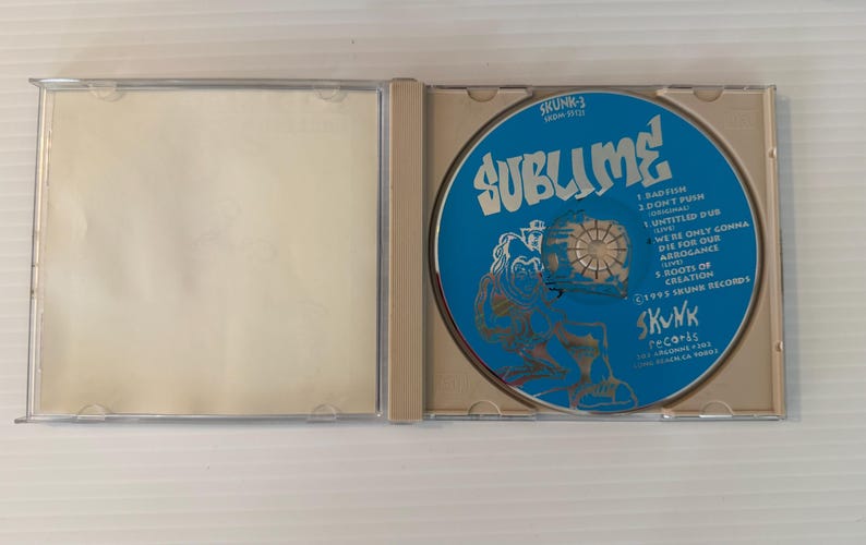 May include: An open CD case with a blue disc featuring the band name "Sublime" in white text. The disc also lists song titles and the "Skunk Records" logo. The case is clear plastic.