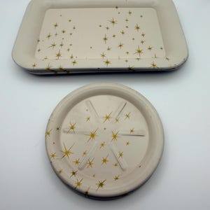 May include: Two cream-colored trays and a coaster with a gold starburst pattern. The rectangular tray has rounded corners. The coaster is round with six grooves and a starburst design. The trays and coaster are stacked.