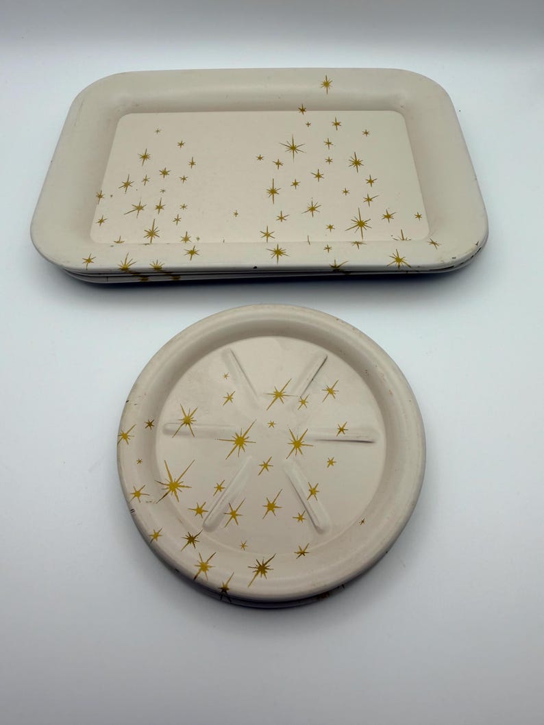 May include: Two cream-colored trays and a coaster with a gold starburst pattern. The rectangular tray has rounded corners. The coaster is round with six grooves and a starburst design. The trays and coaster are stacked.