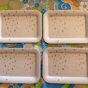 May include: Four rectangular white trays with rounded corners. Each tray has a cream-colored interior decorated with gold starburst patterns. The trays are arranged on a colorful floral patterned surface.