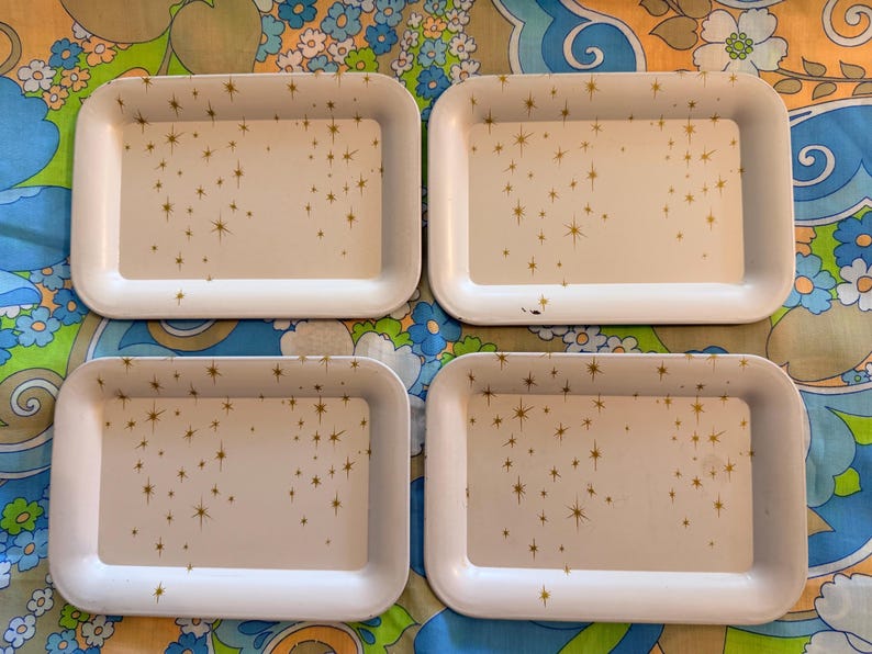 May include: Four rectangular white trays with rounded corners. Each tray has a cream-colored interior decorated with gold starburst patterns. The trays are arranged on a colorful floral patterned surface.