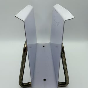 May include: A white metal object with a V-shaped design, supported by two curved metal legs. The object has a rectangular base and angled sides, with a few small holes visible. The metal legs are a darker color than the white body.