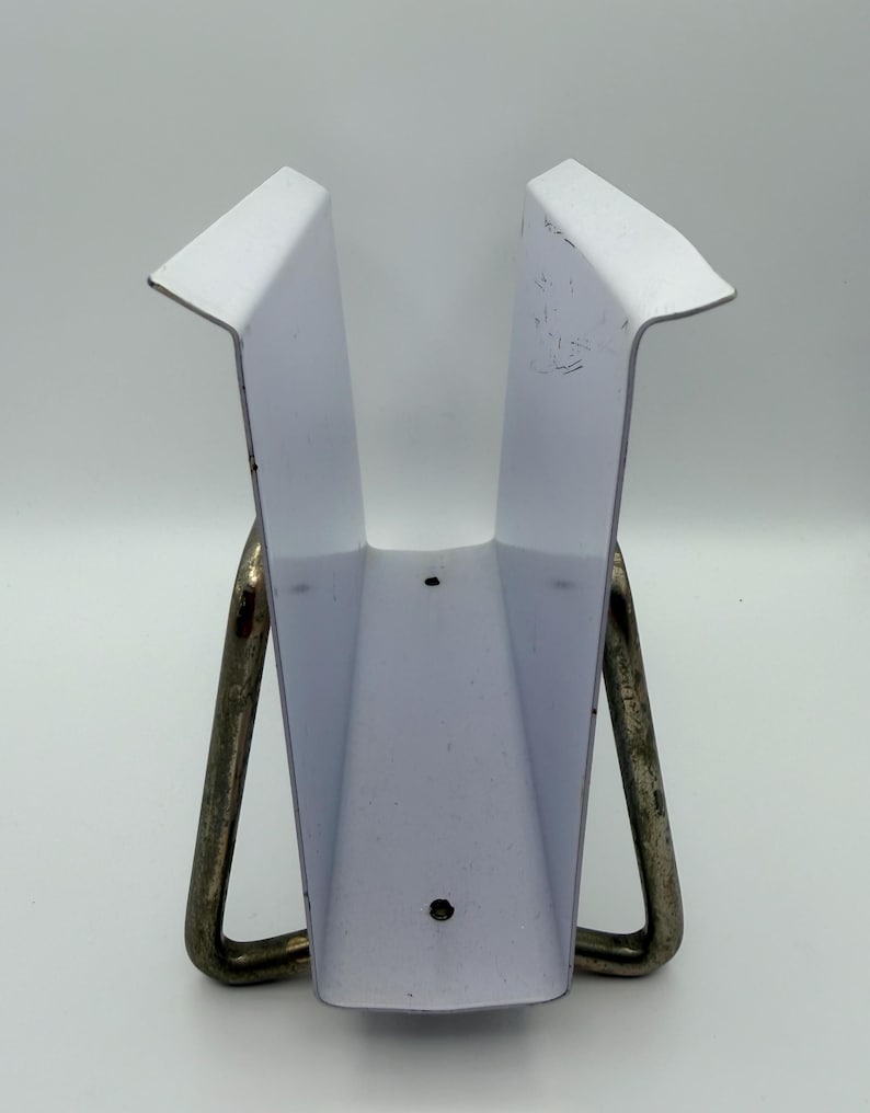 May include: A white metal object with a V-shaped design, supported by two curved metal legs. The object has a rectangular base and angled sides, with a few small holes visible. The metal legs are a darker color than the white body.