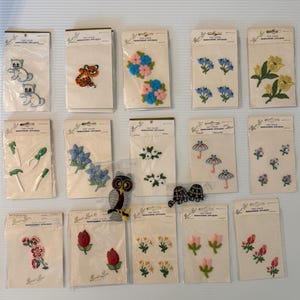 May include: A collection of embroidered appliques in various designs, including flowers, animals, and gardening tools. Each applique is packaged on a card with the text "Fast Color Embroidered Appliques". The colors include blue, pink, yellow, and green.