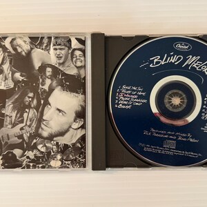 May include: A CD case is open, revealing a CD and its cover art. The CD is dark blue with white text, including the band name "Blind Melon." The cover art is a black and white collage of band members.