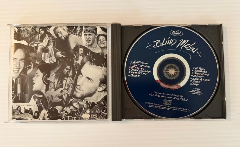 May include: A CD case is open, revealing a CD and its cover art. The CD is dark blue with white text, including the band name "Blind Melon." The cover art is a black and white collage of band members.
