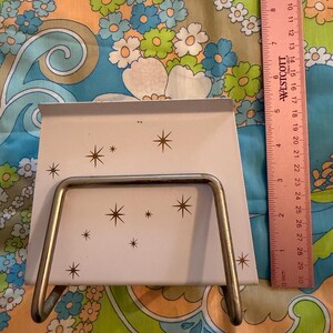 May include: A vintage, off-white metal napkin holder with a starburst pattern. The holder has a silver-colored wire frame and is approximately 6 inches wide. A pink ruler is visible in the background.