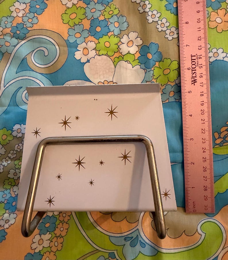 May include: A vintage, off-white metal napkin holder with a starburst pattern. The holder has a silver-colored wire frame and is approximately 6 inches wide. A pink ruler is visible in the background.