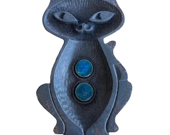 May include: A vintage black cat-shaped ashtray with two circular teal-colored insets. The ashtray has a whimsical design with a smiling face and stylized features. The metal has a slightly weathered appearance, adding to its vintage charm.