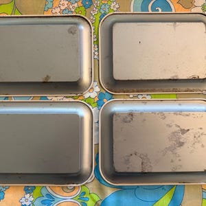 May include: Four rectangular metal trays with rounded corners. The trays have a light gray interior and a cream-colored rim. The trays show signs of wear and tear, with some discoloration and scratches. The trays are arranged in a square formation.