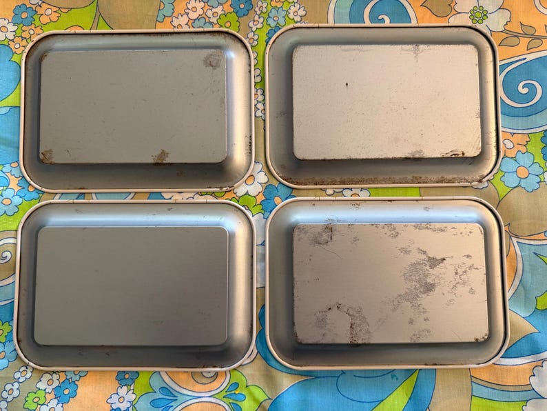 May include: Four rectangular metal trays with rounded corners. The trays have a light gray interior and a cream-colored rim. The trays show signs of wear and tear, with some discoloration and scratches. The trays are arranged in a square formation.