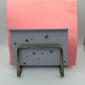 May include: A vintage napkin holder with a light blue front panel decorated with brown starbursts. The holder has a silver metal frame and holds a stack of pink napkins. A retro-style kitchen accessory.