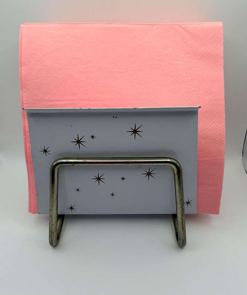 May include: A vintage napkin holder with a light blue front panel decorated with brown starbursts. The holder has a silver metal frame and holds a stack of pink napkins. A retro-style kitchen accessory.
