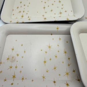May include: A set of off-white rectangular trays with rounded corners. Each tray is decorated with a gold starburst pattern. The trays have a slightly raised edge and appear to be made of metal.