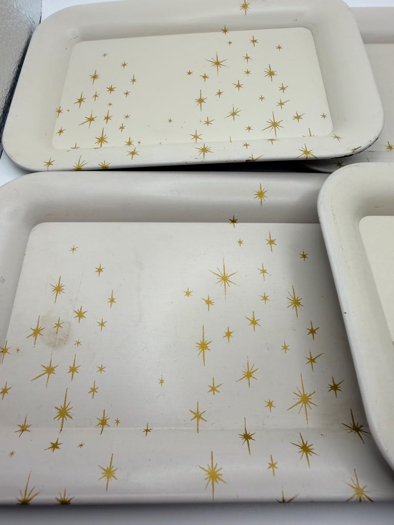 May include: A set of off-white rectangular trays with rounded corners. Each tray is decorated with a gold starburst pattern. The trays have a slightly raised edge and appear to be made of metal.
